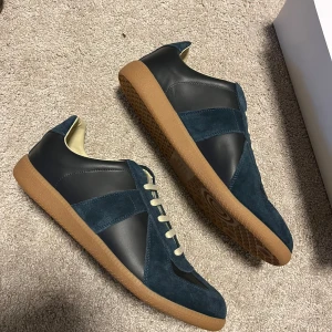 Maison Margiela Gats sneakers blå - Maison Margiela Gatss neakers i mörkblå mocka och svart kalvskinn med beige snörning och gummisula. Klassisk low-top siluett med stilren design och lyxiga materialval. Perfekta för dig som gillar minimalistisk och tidlös stil. Skickas samma dag och priset kan diskuteras vid snabb affär.