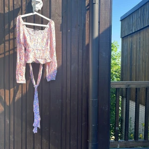 Blommig omlottopp med snöre  - Supersöt blommig topp med långa ärmar och omlottdetalj som knyts framtill. Toppen har volangkant nedtill och smockad rygg för extra skön passform. Färgerna går i rosa, lila och blått på ljus botten. Perfekt för vår och sommar!