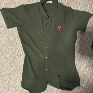 Grön kortärmad skjorta från Ami - Stilren grön skjorta från Ami Alexandre Mattiussi med korta ärmar och klassisk krage. Skjortan har knäppning framtill och en röd broderad logga med hjärta och bokstaven A på bröstet. Perfekt för dig som gillar minimalistisk design med en twist.