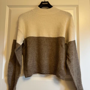 Vit beige/brun höst vinter stickad tröja H&M - Mysig stickad tröja från H&M i tvåfärgad design med vit upptill och beige/brun nedtill. Tröjan har lång ärm, ribbade muddar och en något högre hals. Perfekt för lager-på-lager under kyliga dagar.