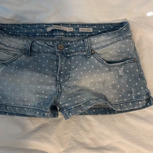 Lågmidjade blå prickiga jeansshorts  - Snygga blå super lågmidjade jeansshorts från Reserved med vita prickar och slitna detaljer. Klassisk femficksmodell med knapp och dragkedja framtill. Perfekta för varma dagar när du vill ha en cool och avslappnad look.