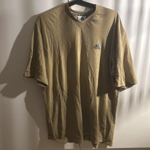 T-shirt från Adidas - Snygg brun/beige t-shirt från Adidas med v ringning, däremot ganska lång krage. Tillverkad i mjuk bomull och bekväm. ‼️Adidas loggan inuti tröjan vid lappen håller på att släppa lite men inget som märks:) 