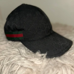 Svart keps från Gucci med logomönster - Snygg svart keps från Gucci med diskret GG-logomönster i tyget och klassiska gröna och röda ränder på sidan. Justerbar rem baktill i skinn. Tillverkad i polyester, bomull och polyamid. Perfekt accessoar för en trendig look.