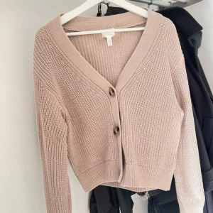 Beige stickad kofta med knappar - Superfin beige stickad kofta med två stora knappar framtill och v-ringad hals. Koftan har lång ärm och en lite croppad passform som ger en trendig look. Perfekt att slänga över en topp för en chill och snygg outfit.