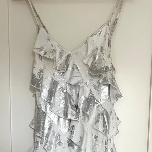Glittrig silvervit volangklänning - Supersnygg lång klänning med tunna axelband och flera lager volanger. Klänningen är vit med glittriga silvriga detaljer som ger en riktigt festlig look. Perfekt för dig som vill sticka ut och glänsa! Materialet känns lätt och skimrande.