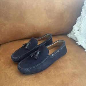 Mörkblå mockaloafers från Massimo Dutti - Snygga mörkblå loafers i mocka från Massimo Dutti med dekorativa tofsar framtill. Skorna har en klassisk rund tå, kontrastsömmar och en gummisula med nitar för extra grepp. Perfekta för en stilren och avslappnad look.