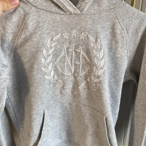 Grå hoodie från Gant med brodyr - Säljer en ljusgrå hoodie från Gant med broderad logga och emblem framtill. Tröjan har huva, känguruficka och är tillverkad i mjuk bomullsmix. Perfekt för chill dagar och snygg till jeans eller mjukisbyxor.