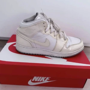 Nike Air Jordan 1 Mid i vitt - Nike Air Jordan 1 Mid i helvitt skinn med klassisk siluett och snörning. Swoosh-logga på sidan och diskret Air Jordan-märke vid ankeln. Perfekta sneakers för dig som gillar stilrena och ikoniska modeller.