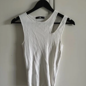 Vit ribbad linnetopp med cut out - Säljer en vit ribbad linnetopp  med cool asymmetrisk cut out vid axeln. Toppen är ärmlös och har en tight passform som sitter snyggt på kroppen. Perfekt för dig som gillar stilrena men ändå unika detaljer.