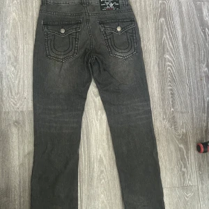 True Religion svarta jeans Joey Super T - Svarta jeans från True Religion, modell Joey Super T. Klassiska kontrastsömmar och stora ficklock bak med tryckknappar. Raka ben och cool faded effekt framtill. Tillverkade i USA i robust denim. 