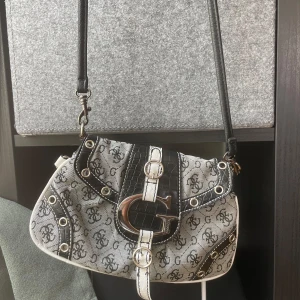 Guess axelväska - Snygg axelväska från Guess med svart och vit monogramdesign, stora silvriga metalldetaljer och croco-imitation på läderdetaljerna. Väskan har justerbar axelrem, dragkedja bak och ikonisk G-spänne framtill. Perfekt för dig som gillar statement-accessoarer.
