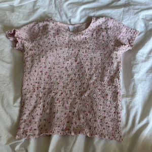 Rosa blommig topp från Zara - Supersöt rosa topp från Zara med små röda blommor och gröna blad. Smockad struktur över hela toppen och volangkant vid ärmslut och nederkant. Kortärmad och stretchig, perfekt för en somrig look.