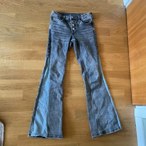 Grå bootcut jeans från H&M, stl 140  - Snygga grå bootcut jeans från H&M med låg midja och justerbar midja. Jeansen har en  egen gjort ljusblå panel längs utsidan av benen och knäppning med flera knappar framtill. Tillverkade i klassiskt denimtyg.