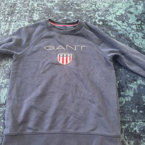 Mörkblå sweatshirt från GANT - Säljer en mörkblå sweatshirt från GANT med broderad logga och sköld på bröstet. Tröjan har rund halsringning, långa ärmar och klassisk passform. Perfekt för dig som gillar stilren och sportig look.