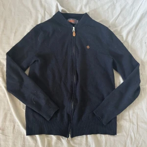 Marinblå  merinoullskofta från Morris - Kofta från Morris i 100% merinoull, marinblå med bronsfärgad broderad logga på bröstet. Stängs med dragkedja och har ribbade muddar vid ärmslut och nederkant. Perfekt för lager-på-lager och snygg till jeans eller chinos. Storlek S men den har krympt så den är storlek xs skulle jag säga, om du vill ha mer bilder är de bara skriva och priset är alltid diskuterbart 