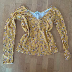 Gul blommig långärmad topp Hollister - Superfin gul långärmad topp från Hollister med blommigt mönster i rosa och grönt. Toppen har v-ringning och rynkdetalj framtill, vilket ger en snygg passform. Perfekt för dig som gillar färg och vill sticka ut lite extra.