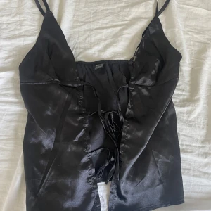Svart satinlinnetopp från SHEIN - Säljer en svart glansig linne-topp från SHEIN i satinliknande material. Toppen har smala axelband och snygg snörning framtill. Väldigt bra skick och super fin till sommaren ❤️
