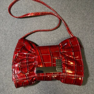 Röd axelväska från Fiorelli - Säljer en röd axelväska från Fiorelli i snygg rosettformad design. Väskan har ett metallspänne framtill, justerbar axelrem och flera fack inuti, inklusive dragkedjefack. Storlek: ca 24x15x5