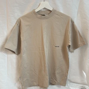 Beige t-shirt från Yoke - Säljer en stilren beige t-shirt från Yoke med liten svart textdetalj på bröstet. T-shirten har klassisk passform, rund halsringning och är tillverkad i mjuk bomull.