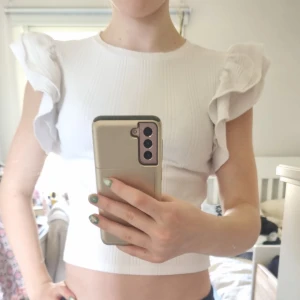 Vit ribbad topp med volangärmar från Gina Tricot - Säljer en söt vit ribbad topp från Gina Tricot med korta volangärmar. Toppen har en rund halsringning och är figurnära i modellen. Perfekt för dig som gillar en feminin och trendig stil!Har användit några gånger men i nyskick.  Frakt ingår inte. Säljer för att jag har växt ur dem. 