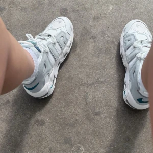 Nike Air More Uptempo vita sneakers - Säljer ett par vita Nike Air More Uptempo sneakers med stora grå bokstäver på sidorna och blå detaljer. Skorna har snörning och en robust, sportig design med tydlig Nike-logga på sulan. Perfekta för dig som gillar streetwear och unika sneakers. Jag har knappt använt dem. Skokartongen finns med! 