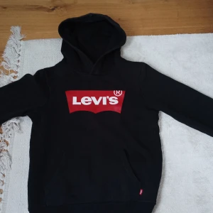 Svart hoodie från Levi's med röd logga - Svart hoodie från Levi's med stor röd och vit logga på bröstet. Klassisk huva och känguruficka framtill. Perfekt för en avslappnad och cool stil.