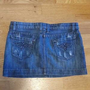 Jeanskjol från Life Love Fashion - Säljer en y2k klassisk blå jeanskjol med broderade bakfickor  Kjolen har en rak passform och är kort i modellen (minikjol) Snygga slitningar och detaljer på fickorna.
