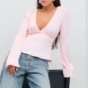 Ljusrosa omlottopp med v-ringning - Supersöt ljusrosa omlottopp med djup v-ringning och långa ärmar. Toppen har en knytrosett i ryggen som ger en fin detalj. Perfekt att styla med jeans eller kjol för en trendig look.