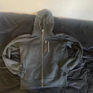 Svart hoodie med dragkedja från Equipe Sports - Svart hoodie med hel dragkedja framtill och justerbar huva. Bröstficka med dragkedja och broderad logga på vänster sida. Perfekt för träning eller chill. Klassisk passform och mjukt material.