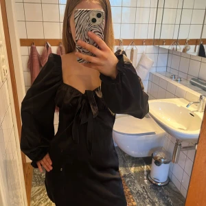 Svart klänning med puffärmar och rosett - Säljer en svart kortklänning med långa puffärmar och fyrkantig urringning. Klänningen har en rosett framtill och är i satin. 