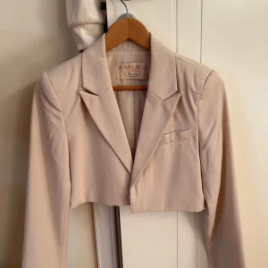Beige croppad kavaj från Missguided - Säljer en beige croppad kavaj från Missguided med klassisk krage och lång ärm. Kavajen har en stilren design med en liten bröstficka och dragkedja vid ärmslutet. Perfekt för en trendig och stilren look.
