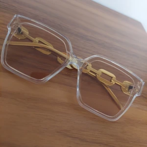 Transparenta fyrkantiga solglasögon med gulddetaljer - Stilrena solglasögon med transparent båge och fyrkantiga linser. Snygga guldfärgade kedjedetaljer längs skalmarna ger en unik och trendig look. Perfekta för dig som vill sticka ut med accessoarer i guld och klara färger.