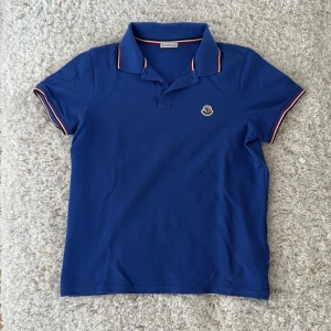 Blå pikétröja från Moncler - Säljer en blå pikétröja från Moncler med klassisk krage och knappslå. Tröjan har Moncler-logga på bröstet och röd-vita detaljer på krage och ärmslut. Använd ett fåtal gånger. Säljes pga att den är för liten. 
