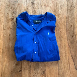Ralph Lauren – Koboltblå skjorta i bomull - Klassisk långärmad skjorta i 100% tvålagers bomull med limegrön broderad logga. Snygg och stilren skjorta med button-down-krage och tidlös preppy-känsla.  Storlek: XL (rätt liten i storleken, skulle säga att den passar M - L)  Färg: Koboltblå med limegrön logga Material: 100% bomull (two-ply cotton) Skick: Mycket fint skick – inga fläckar eller hål Stil: Preppy / Klassisk / Vintage Passform: Rak modell med button-down-krage Nypris: ca 1 300 kr Pris: 250 kr  Frakt: Skickas snabbt & spårbart 