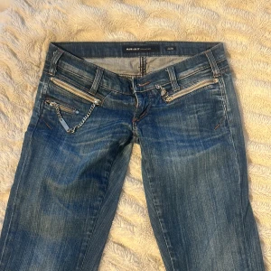 Blå Miss Sixty jeans med kedjedetalj - Säljer ett par blå jeans från Miss Sixty med snygga dragkedjor och kedjedetalj framtill. Jeansen har ich raka ben och klassiska bakfickor med dekorativa sömmar. Perfekta för dig som gillar en cool och unik stil.