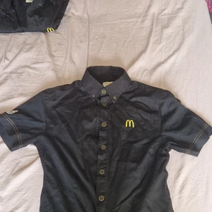 Svart kortärmad skjorta från McDonald's - Svart skjorta med korta ärmar och klassisk krage från McDonald's Professional Workwear Edition. Skjortan har knappar framtill och den ikoniska gula McDonald's-loggan broderad på bröstet. Perfekt för dig som gillar unika plagg med streetkänsla.