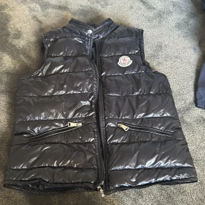 Svart  Moncler gui väst - Säljer en svart glansig dunväst från Moncler med ståkrage och dragkedja framtill. Västen har två snedställda fickor med dragkedja och Moncler-logga på bröstet. Perfekt för lager-på-lager och stilren look.