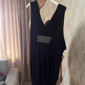 Svart långklänning med strassdetalj - Säljer en svart långklänning med djup v-ringning både fram och bak. Klänningen har en snygg strassdetalj framtill och knytband i ryggen. Perfekt för dig som vill ha en elegant och stilren look.