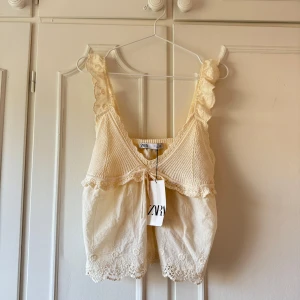 Krämfärgad/ljusgul topp från Zara  - Supersöt beige linnetopp från Zara med virkade axelband och spetsdetaljer nertill. Toppen har v-ringning, knyt framtill och är ärmlös. Perfekt för varma dagar och ger en romantisk känsla till outfiten.