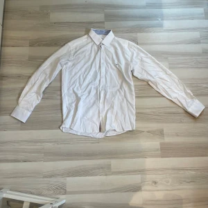 Vit skjorta från Tommy Hilfiger - Säljer en klassisk vit skjorta från Tommy Hilfiger i storlek L. Skjortan har blå- och vitrandig insida på kragen och detaljer vid knappslån. Långärmad modell med knappar och diskret logga på bröstet. FUNKAR UTMÄRKT TILL M!