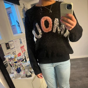 Svart stickad tröja med text från Chica London - Säljer en svart stickad tröja från Chica London med stora bokstäver 'AMOUR' i olika material och färger framtill. Tröjan har långa ärmar och ribbade muddar vid ärmslut och nederkant. Perfekt för dig som gillar statement-plagg!