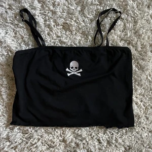 Svart tubtopp med dödskalle från Current Mood - Cool svart tubtopp från Current Mood med smala justerbara axelband och ett broderat dödskallemotiv framtill. Toppen är tillverkad i bomull och spandex för en bekväm och stretchig passform. Perfekt för dig som gillar edgy stil!