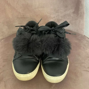 Svarta sneakers med fuskpäls och rosett - Svarta sneakers med stora svarta rosetter och fluffiga fuskpälsdetaljer framtill. Skorna har vit platt sula och dragkedja på sidan. Perfekta för dig som vill sticka ut med din stil.💗