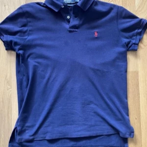 Mörkblå pikétröja från Polo Ralph Lauren - Säljer en klassisk mörkblå pikétröja från Polo Ralph Lauren med röd broderad logga på bröstet. Tröjan har krage, knappar vid halsen och är kortärmad. Perfekt för en stilren och avslappnad look nu till sommaren! Den har inte defekter och nypris är runt 1200💕