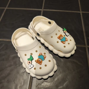 Vita sandaler med nallebjörnsmotiv - Säljer ett par vita sandaler i syntetmaterial med bruna prickar och söta nallebjörnsdekorationer. Skorna har en vågig sula och är dekorerade med texten 'POW' i grönt och vitt. Perfekta för dig som gillar lekfull stil.