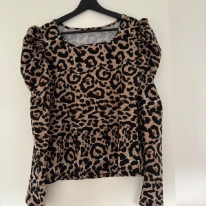 Leopardmönstrad topp med puffärm - Säljer en beige och svart leopardmönstrad topp med långa ärmar och markerade puffärmar. Toppen har rund halsringning och är i ett glansigt material som ger en cool look. Perfekt för dig som vill sticka ut lite extra. Storlek M. Aldrig använd endast testad 