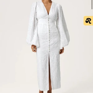 Vit Positano långklänning med puffärm och knappar - Säljer en vit långklänning i Positano-stil med v-ringning, långa puffärmar och dekorativa knappar framtill. Klänningen har en slits framtill och ett subtilt blommigt mönster i tyget. Perfekt för sommarens speciella tillfällen.