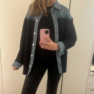 Sandro jacka - Säljer en oversized svart overshirt med blå jeansdetaljer på axlar, krage och ärmslut. Jackan har knappar framtill och en avslappnad passform.