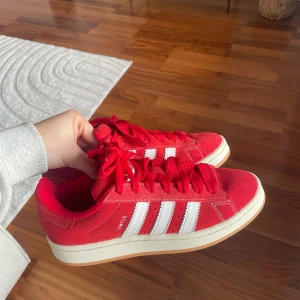 Röda Adidas Campus sneakers - Röda Adidas Campus sneakers med vita ränder och vit sula. Skorna har klassisk design med snörning och ovandel i mocka. Kommer inte till användning, har endast använt 2 gånger. Kartong ingår ej 