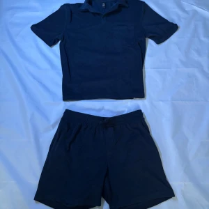 Mörkblå frotté T-shirt med shorts - Säljer en mörkblå frotté t-shirt med bröstficka. T-shirten och shortsen har en avslappnad passform och är tillverkad i ett mjukt material som känns skönt mot huden. Perfekt för en stilren och enkel look för sommaren. Näst intill aldrig använd.